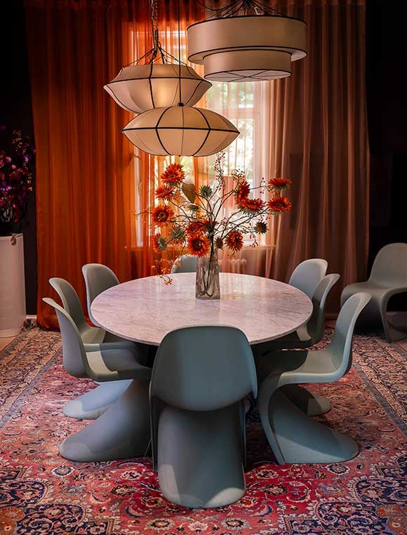 Färgglad interiör av ett vardagsrum med bord och stolar. Stor vase med röda blommor på. Beige lampor med mysigt ljus