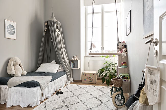 Barnrum med gråmålade väggar och trägolv, med en rutig matta i färgerna vit och svart. I hörnan står en säng med grå sänghimmel, och mot väggen står en bokhylla i ljust trä med leksaker ovanpå.