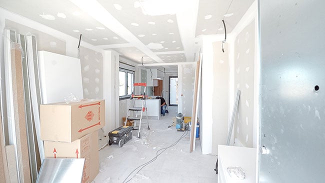 Nyproduktion av bostad i Spanien, bygget sker inomhus. Rummet har gråa väggar med vita spackelfläckar, på golvet står flyttkartonger, en stege, byggnadsmaterial och verktyg.