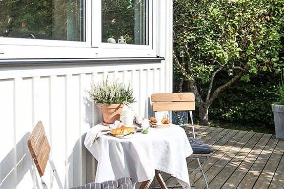checklista vid visning - uteplats med dukat bord med frukost eller fika, solen lyser och uteplatsen är omgiven av gröna gräsmattor och lummiga träd och buskar